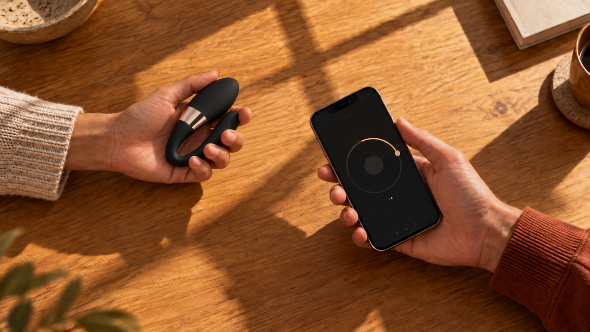 Deux mains sur une table en bois : l'une tient un vibromasseur de couple noir en forme de C, l'autre un smartphone affichant l'application de pilotage