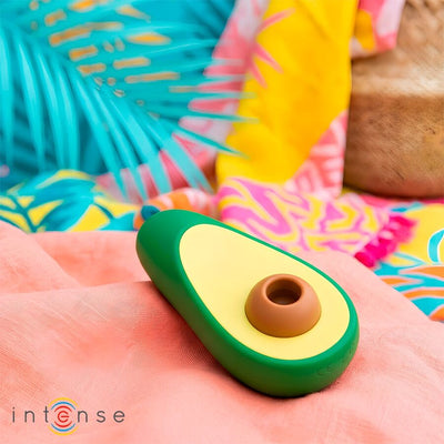Stimulateur vert Intense Waki en forme d’avocat, posé sur un tissu rose parmi des coussins colorés